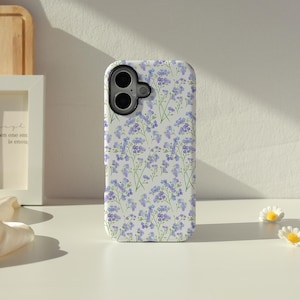 Op de afbeelding: Een witte smartphonehoes met een herhalend patroon van kleine, lichtpaarse vergeet-mij-nietjes met groene stengels en bladeren. De hoes heeft een zwarte uitsparing voor de cameralens in de linkerbovenhoek voor een dubbele camera.
