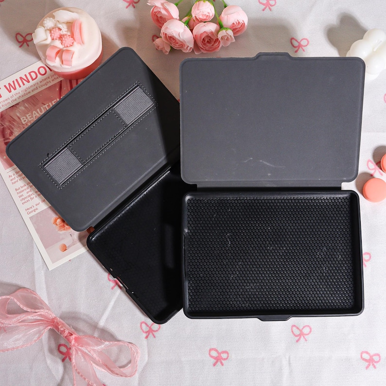 May include: A set of three open, dark gray tablet cases. One case is open, revealing a textured black interior. Another case is open, displaying a gray elastic strap. The cases are on a light pink surface with pink bow accents and flowers.