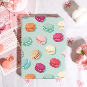 May include: A light blue tablet case featuring a pattern of colorful macarons. The macarons are in shades of orange, pink, green, beige, and purple. The case is rectangular and has rounded corners. The background is a light blue color.