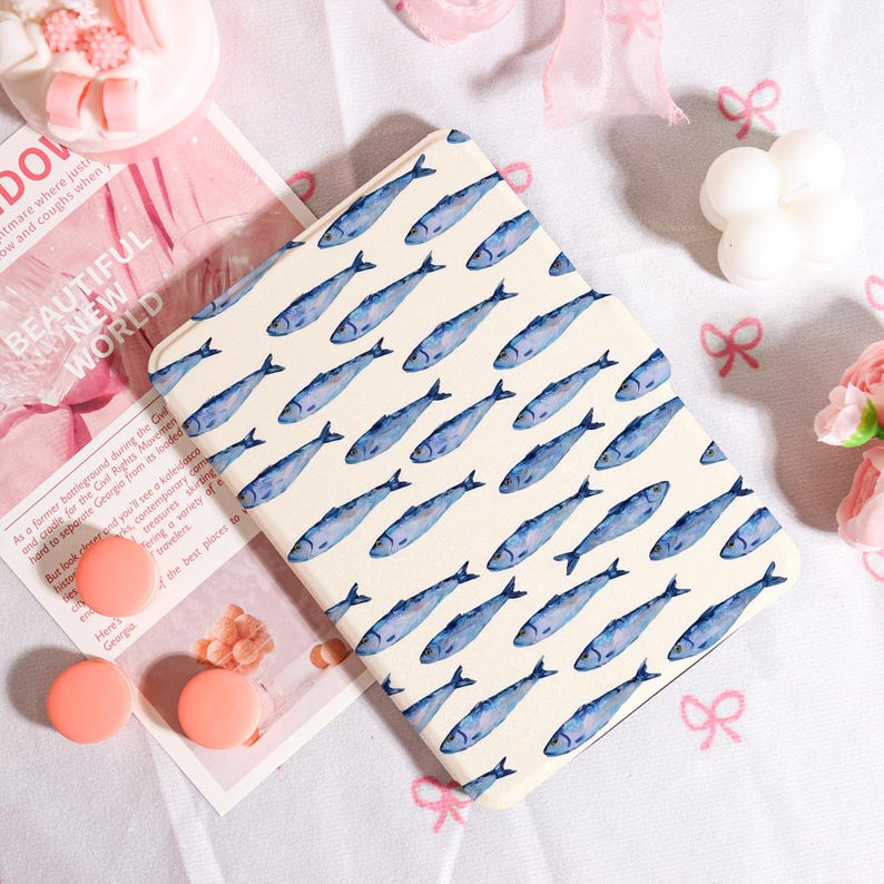 May include: A cream-colored notebook featuring a repeating pattern of blue watercolor fish. The notebook is rectangular and lies on a pink surface with other decorative items, including a pink bow and a small candle.