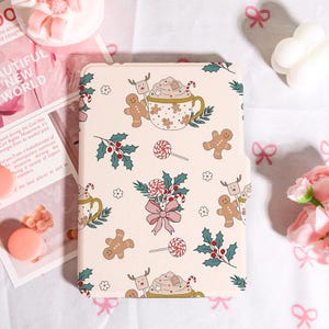 Könnte beinhalten: Eine hellrosa Tablet-Hülle mit einem sich wiederholenden Muster aus Lebkuchenmännern, Stechpalmen, Lollis und Kaffeetassen. Das Design umfasst eine rosa Schleife und kleine weiße Blumen. Die Hülle hat ein festliches, weihnachtliches Thema.