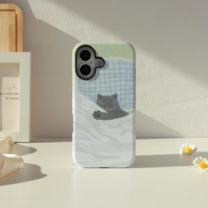 May include: A phone case featuring a gray cat sleeping under a white blanket. The case has a light green top and a blue plaid pattern. The cat is the focal point, with its eyes closed and paws visible.