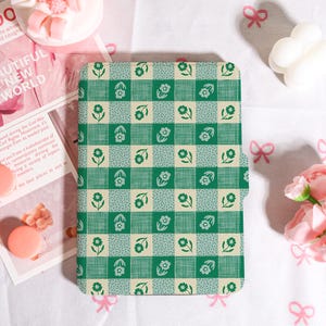 May include: A green and cream tablet case with a floral and checkered pattern. The case has a repeating design of green flowers and squares. The rectangular case appears to be made of a durable material.