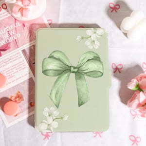 May include: A pale green tablet case with a large watercolour bow in a matching shade. White floral accents are placed around the bow. The case has a smooth finish and a rectangular shape. The background includes a magazine and decorative items.