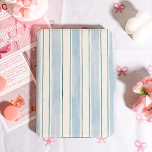 Puede incluir: Una funda rectangular con un patrón de rayas verticales en azul claro y blanco. La funda tiene una estética suave, similar a la acuarela. El fondo incluye flores rosas, una revista y elementos decorativos.
