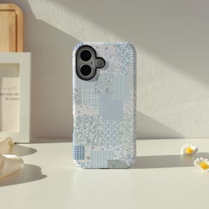 Peut inclure: Une coque de smartphone avec un motif patchwork bleu clair et blanc. Le design se compose de divers carrés présentant des motifs floraux, de fines rayures verticales et de petits carreaux, créant un collage cohérent de tons bleu clair.