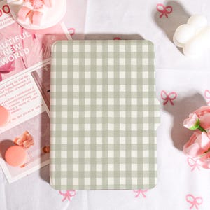 Puede incluir: Una funda rectangular con un patrón de cuadros de color verde claro y blanco. La funda está cerrada y tiene una superficie lisa. El fondo incluye una revista con el texto "BEAUTIFUL NEW WORLD".
