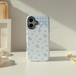 Puede incluir: Una funda de teléfono azul claro que presenta una sección superior con un patrón de cuadros vichy blanco y azul, un gráfico de lazo azul claro central y una sección inferior decorada con un patrón de pequeñas rosas azules sobre fondo blanco.