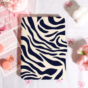 May include: A rectangular case with a cream-colored background and a navy blue zebra stripe pattern. The case is likely for a tablet or e-reader. The case is on a white surface with pink bows and flowers.