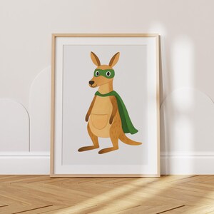 Puede incluir: Una impresión enmarcada con un superhéroe canguro de dibujos animados. El canguro está representado en tonos marrones y beige, con una capa y máscara verdes. La obra de arte está sobre un fondo gris claro, enmarcada en madera natural.