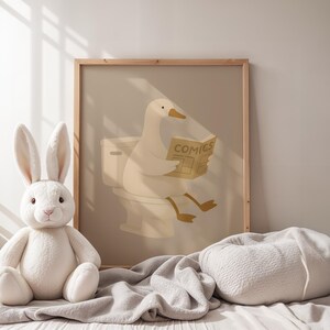 Neutral nursery wall art print, soft beige duck bathroom poster, minimalist children's room decor, gentle animal illustration art, calm nursery bathroom sign, Scandinavian baby room print, warm neutral kids wall art, peaceful nursery poster, duck reading newspaper illustration, natural wood frame nursery art, monochromatic baby room decor, hygge children's wall hanging, serene kids bathroom art, gender neutral nursery print, modern minimalist baby decor, soft color palette children's art.