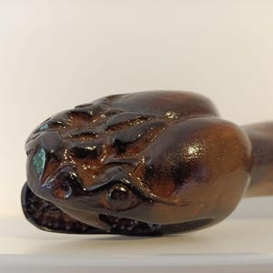 May include: A close-up of a wooden sculpture of a creature's head, featuring a glossy, dark brown finish. The sculpture has detailed facial features and is set against a plain white background. The head is the focal point, with the rest of the body extending to the right.