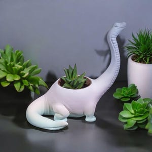 Puede incluir: Un macetero de dinosaurio de cerámica con un degradado de azul claro a blanco. Tiene una planta suculenta dentro. El macetero tiene un cuello y una cola largos.