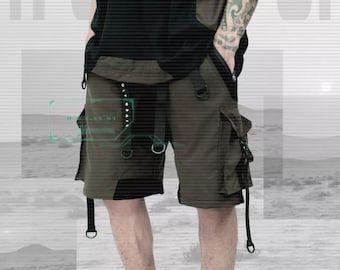 Op de afbeelding: Een persoon draagt een zwarte en groene cargo-short met meerdere banden en gespen. De short wordt gedragen over een zwart T-shirt en de tekst "H.G.SS-01" staat op het T-shirt gedrukt.