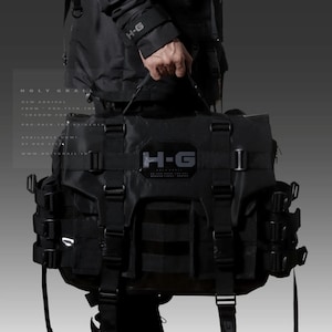 May include: A black, tactical-style messenger bag with multiple straps and buckles. The bag has a large, rectangular shape and features the text "H-G" in white on the front.