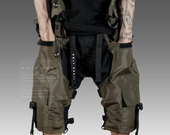 Op de afbeelding: Groene cargo shorts met meerdere banden en gespen. De shorts hebben grote zakken aan de zijkanten en een zwarte riem. De tekst "HOLY GRAIL" is gedrukt op het rechterbeen.
