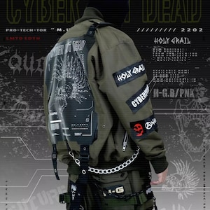 May include: A green bomber jacket with black and white patches, including the text "Cyber Dead" and "Holy Grail". The jacket has a zipper closure and multiple pockets. The model is wearing the jacket with black pants and a black beanie.