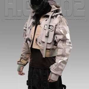 Puede incluir: Una persona que lleva una chaqueta gris y beige con un casco negro y un gráfico en blanco y negro en la manga. La chaqueta tiene varios bolsillos y correas. El texto "H.G.02" es visible en la chaqueta.