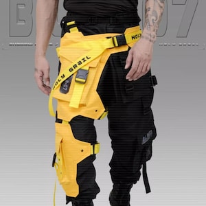 Puede incluir: Pantalones cargo negros con detalles y correas amarillas. Los pantalones tienen m&uacute;ltiples bolsillos y un accesorio amarillo tipo funda en la pierna izquierda. El texto "HOLY GRAIL" es visible en una correa amarilla. El estilo es futurista y urbano.