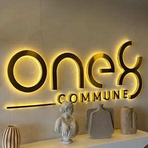 以下が含まれることがあります： 壁に取り付けられた、金色の照明付きサイン。上部に「one8」、下部に「COMMUNE」の文字。白い棚には、胸像や花瓶など、装飾品が置かれています。