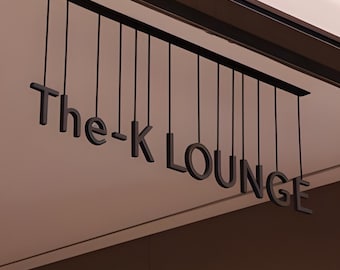 May include: Black letters spelling "The-K LOUNGE" are suspended from a black bar. The letters hang from thin black cords against a light brown ceiling. The sign is angled, with the letters in a sans-serif font.