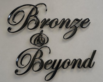 May include: A black wall sign featuring the words Bronze & Beyond in a cursive script font. The letters have a glossy finish and are arranged in two lines with an ampersand symbol centered between the words Bronze and Beyond.