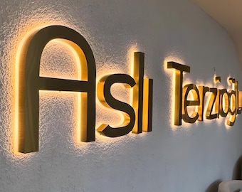 May include: Gold-colored, illuminated sign on a textured white wall. The sign reads "Aslı Terzioğlu" in a stylized font, with the letters casting a warm glow. The sign is mounted on a wall, with a glimpse of seating in the background.