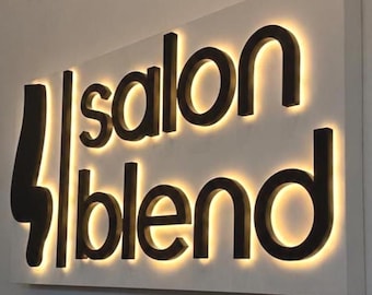 May include: A rectangular wall-mounted sign featuring the black text salon blend in a lowercase sans-serif font. The letters are mounted on a white panel and are backlit with a warm yellow glow that creates a halo effect around each character.