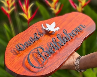 May include: A wooden sign with the words "David's Bethlehem" in black script and a white dove. The sign has a natural wood grain finish and an organic shape. The background features green foliage and red and yellow flowers.