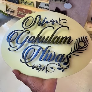 May include: A round, cream-colored sign with the words "Sri Gokulam Nivas" in black and blue script. A black peacock feather design is on the right side. The sign is likely a house nameplate or decorative item.