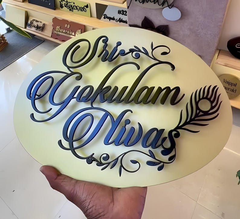 May include: A round, cream-colored sign with the words "Sri Gokulam Nivas" in black and blue script. A black peacock feather design is on the right side. The sign is likely a house nameplate or decorative item.