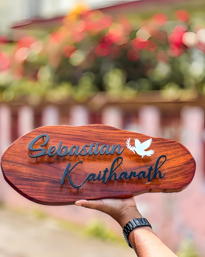 May include: A wooden nameplate with the name "Sebastian Kaitharath" in black script. A white dove with an olive branch is on the right. The nameplate is held up against a blurred background of greenery and flowers.