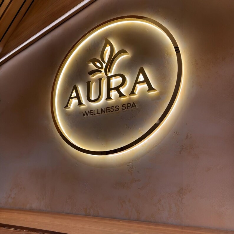 May include: Aura Wellness Spa sign. The sign is circular with a gold border and a glowing light. The word "AURA" is in gold letters, with a leaf design above it. Below the word "AURA" is the text "WELLNESS SPA".