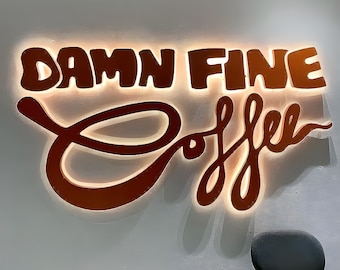 May include: A wall-mounted sign with the words "Damn Fine Coffee" in a stylized, illuminated design. The letters are a warm brown color, with a bright, glowing outline. A dark gray chair is visible below the sign.