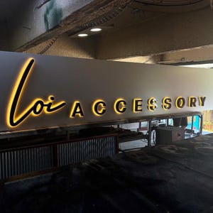 May include: A sign that reads "Loi Accessory" in a modern, illuminated font. The word "Loi" is in a cursive script, and "Accessory" is in a bold, sans-serif font. The letters glow with a warm, golden light.