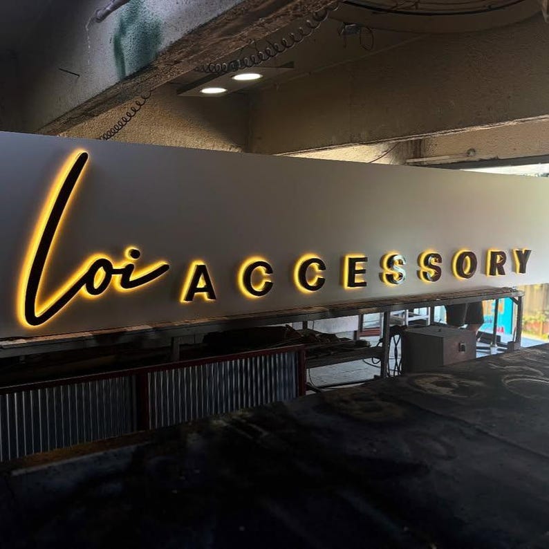 May include: A sign that reads "Loi Accessory" in a modern, illuminated font. The word "Loi" is in a cursive script, and "Accessory" is in a bold, sans-serif font. The letters glow with a warm, golden light.