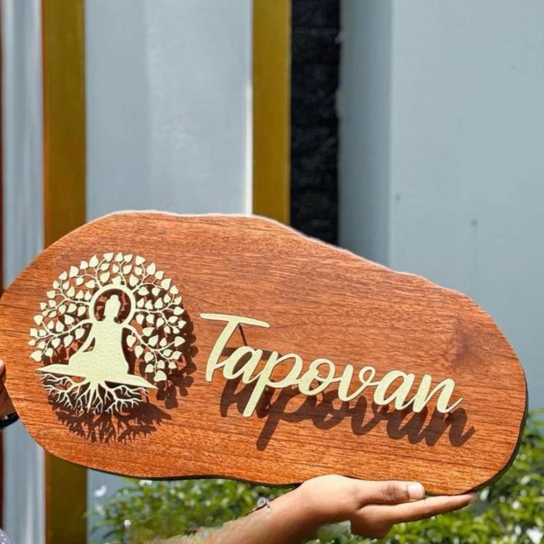 May include: Wooden sign with the name "Tapovan" in a cursive font. The sign features a gold-colored tree of life design with a seated figure. The sign is brown with a natural wood grain.