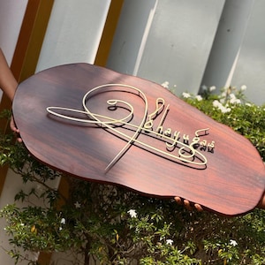 May include: A wooden sign with a dark brown finish and a gold-colored wire design. The sign has the name "Bhayu" in a cursive font. The sign is held in front of a green bush.