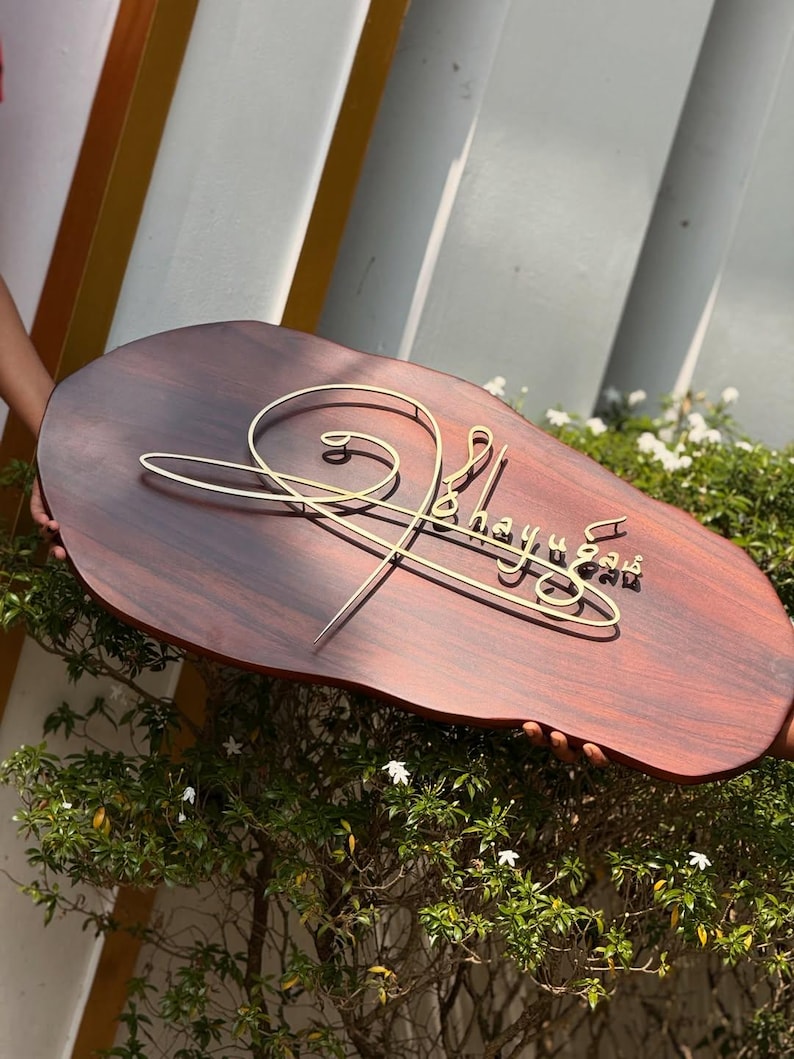 May include: A wooden sign with a dark brown finish and a gold-colored wire design. The sign has the name "Bhayu" in a cursive font. The sign is held in front of a green bush.