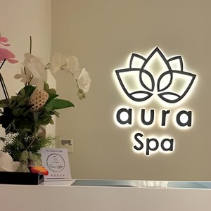 May include: Aura Spa sign with a lotus flower design, illuminated with a soft glow. The words "aura" and "Spa" are below the logo. A floral arrangement and a small sign are visible in the foreground.
