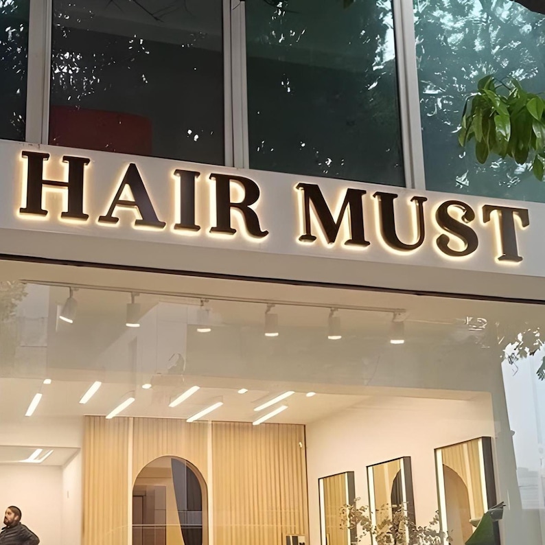 May include: A storefront sign with the words "HAIR MUST" in large, illuminated letters. The sign is mounted above a glass window, with interior lights and decor visible inside. The overall aesthetic suggests a hair salon or beauty establishment.