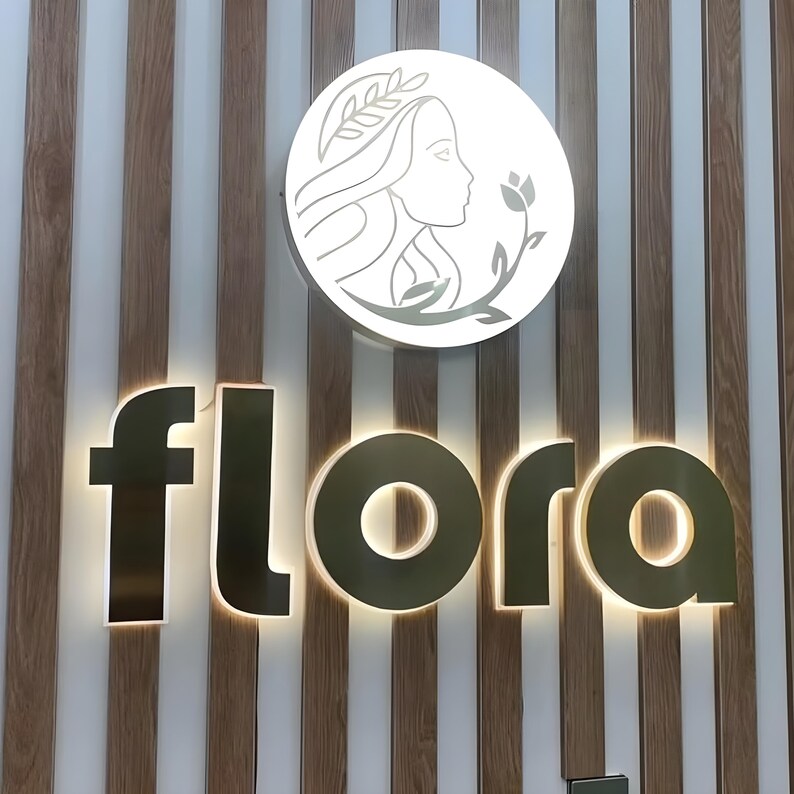 May include: Illuminated sign with the word "flora" in gold lettering. Above, a circular sign features a woman's profile and floral elements. The background has vertical wood and white paneling.