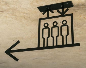 May include: Black metal elevator sign with three figures inside a square, arrows pointing up and down, and a left-pointing arrow. The sign is mounted on a beige textured surface.