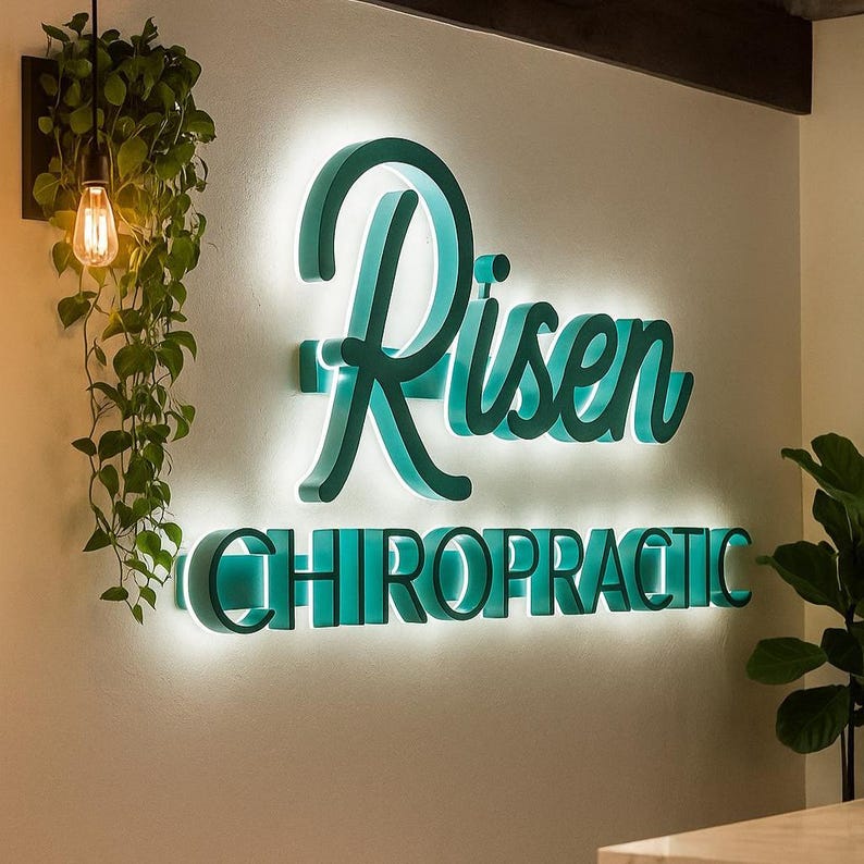 May include: A wall-mounted sign for a chiropractic clinic. The sign features the word "Risen" in a teal script font above the word "CHIROPRACTIC" in teal block letters. Both words are illuminated with a white glow.