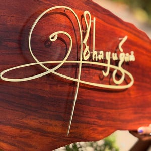 May include: A wooden sign with a rich, reddish-brown finish. The sign features gold-colored, stylized lettering that reads "Ohayugah". The sign has a unique, organic shape, adding to its handcrafted appeal. The sign is likely for home decor.