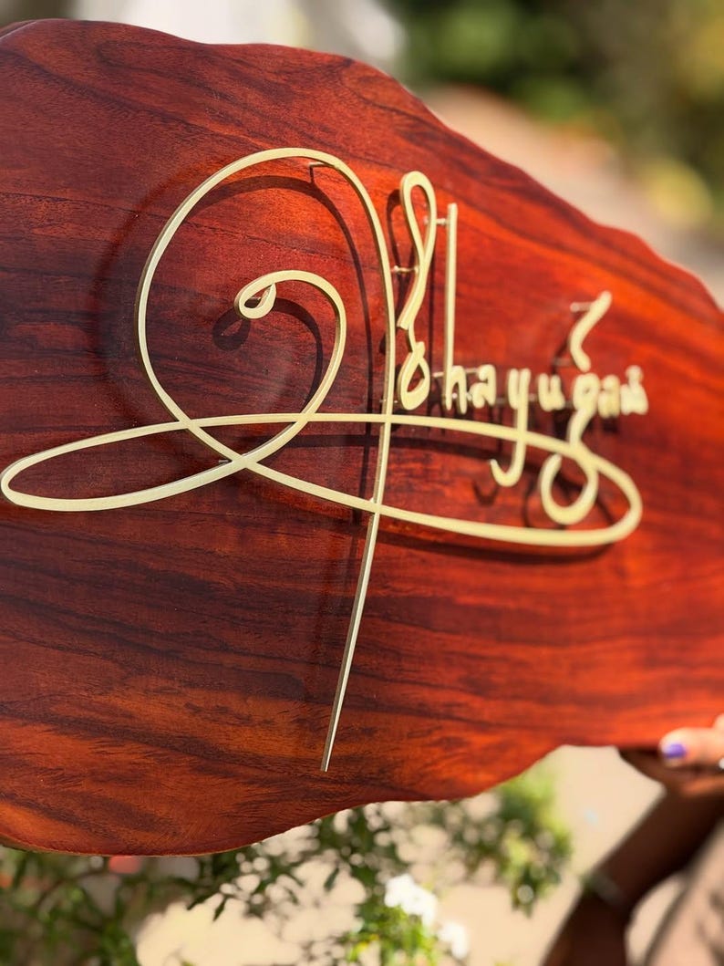May include: A wooden sign with a rich, reddish-brown finish. The sign features gold-colored, stylized lettering that reads "Ohayugah". The sign has a unique, organic shape, adding to its handcrafted appeal. The sign is likely for home decor.