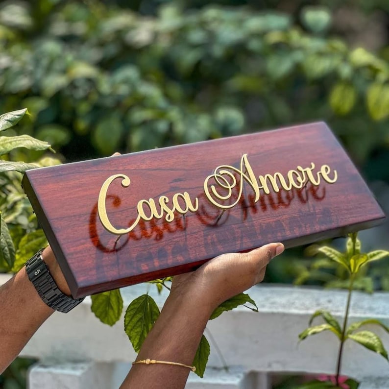 May include: A rectangular wooden sign with the words "Casa Amore" in gold script. The sign is a rich, dark brown color and is being held up by a person. The background is blurred, with green foliage.