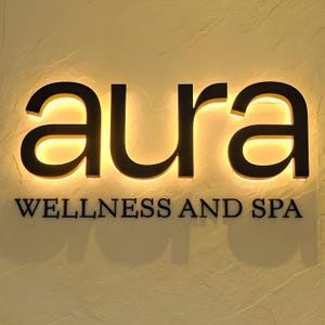May include: A backlit sign that reads "aura" in a bold, black font, with the words "WELLNESS AND SPA" below. The sign is mounted on a textured, light-colored wall, creating a warm, inviting ambiance.
