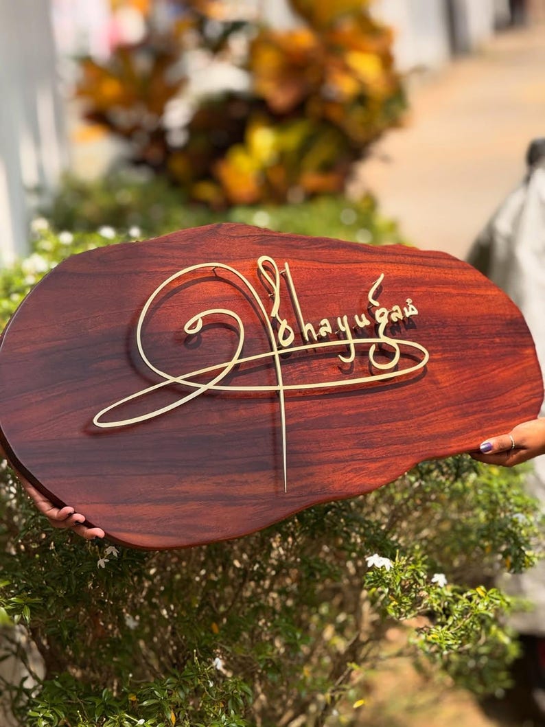May include: A wooden sign with a dark brown finish and a unique, organic shape. The sign features elegant, gold-colored script that reads "Ohayugaia". The sign is held in front of a blurred background of greenery.