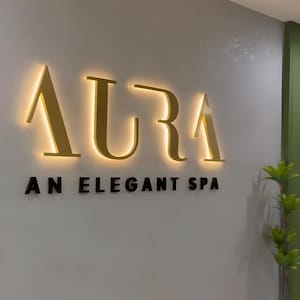 May include: The word "AURA" in gold, illuminated letters is mounted on a light gray wall. Below, the text "AN ELEGANT SPA" is in black letters. A green accent wall and small plant are visible.
